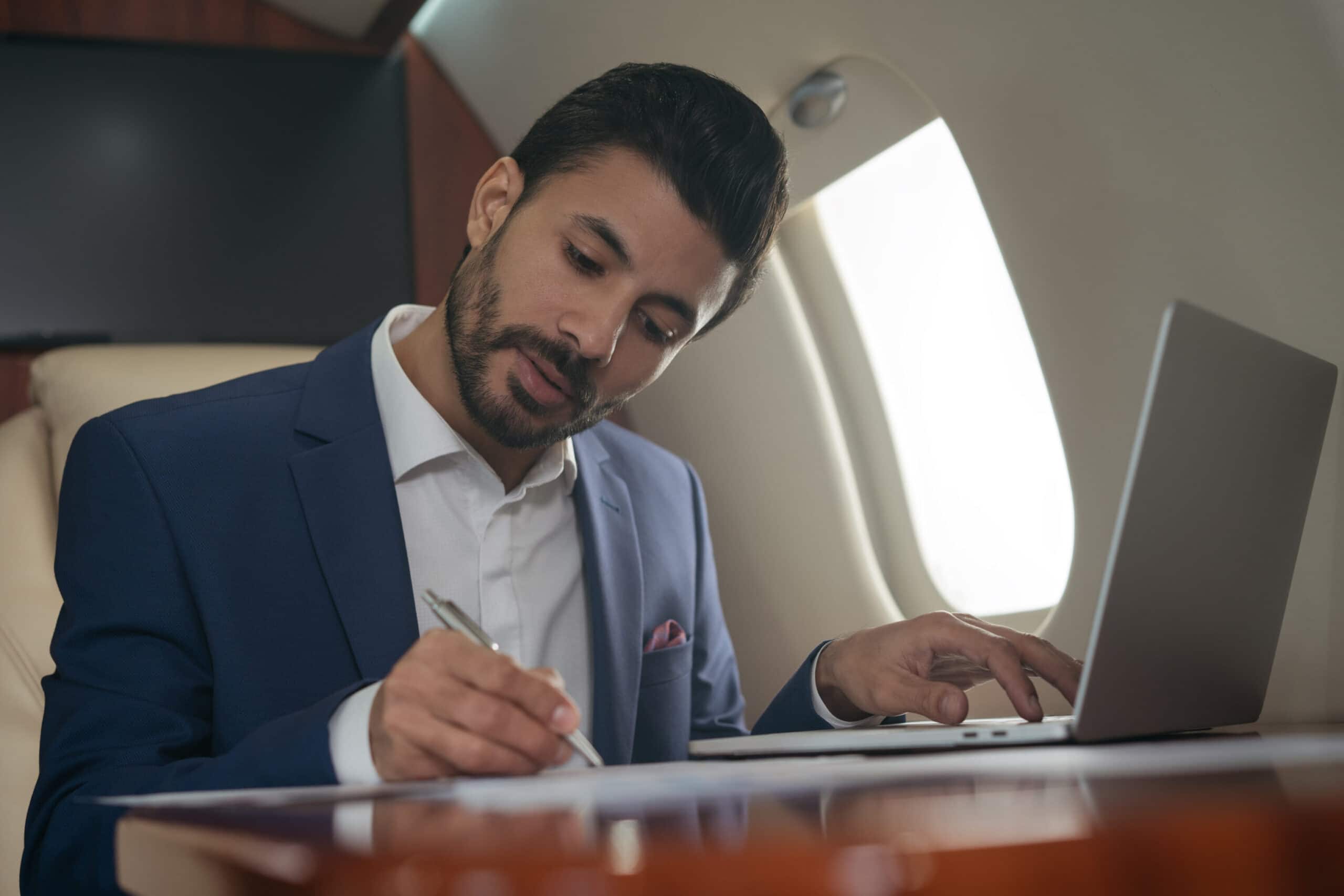 Businessman using laptop computer, typing, working online sitting in airplane. Confident middle eastern entrepreneur taking notes, signs contract flying luxury private jet Business man in private jet